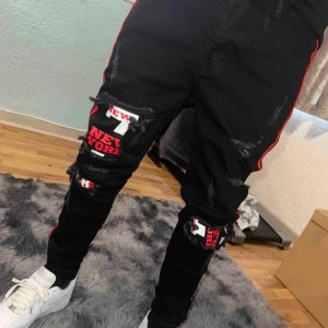 Svarta jeans med röda detaljer - Snygga svarta jeans med slitna detaljer och coola New York-tryck. De har röda ränder längs sidorna som ger en sportig look. Perfekta för dig som vill sticka ut!