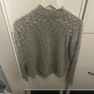 Grå tröja med pärlor från Divided - Säljer en stilren grå tröja från Divided med dekorativa pärlor över axlarna och ärmarna. Tröjan har en normal passform och långa ärmar, perfekt för en elegant look. Passar bra till både jeans och kjol.