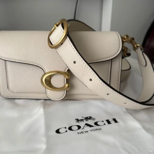 Beige axelväska från Coach - Snygg beige axelväska från Coach i skinn med guldfärgade detaljer. Väskan har en justerbar axelrem och ett stort C-spänne framtill. 