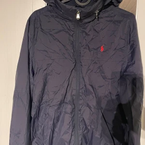 Mörkblå vindjacka från Ralph Lauren - Snygg mörkblå vindjacka från Ralph Lauren med dragkedja och huva. Jackan har en liten röd logotyp på bröstet och är perfekt för blåsiga dagar. Den är lätt och smidig att bära med sig.