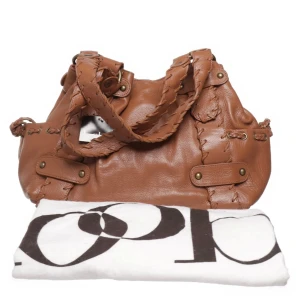 Kooba Sienna  - Oanvänd! Kooba är välkänd Y2K-väska som är populär bland bloggare och kändisar. Snygg brun axelväska i skinn med unika flätade detaljer och nitar. Väskan har en robust design med dubbla handtag och dekorativa sömmar. Perfekt för att ge din outfit en cool touch.