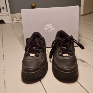 Svarta Nike Air Force 1 - Snygga svarta Nike Air Force 1 sneakers med klassisk design och snörning. Perfekta för en stilren look. Skorna har en robust sula och är tillverkade i syntetmaterial.