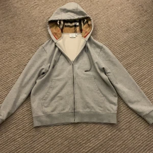 Burberry tröja - Snygg grå hoodie från Burberry med dragkedja och klassiskt rutigt foder i huvan. Använd endast 2 gånger och är i perfekt skick! Perfekt för att utveckla sin klädstil.