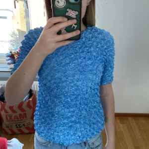 Blå topp med textur - Säljer en unik blå topp med en intressant textur. Toppen har korta ärmar och en normal passform. Perfekt för att ge en färgklick till din garderob.Den hade ett hål i armhålan, men vi har sytt ihop hålet nu. Så det syns knappast.