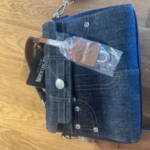 Jeansväska från True Religion - Snygg jeansväska från True Religion med bruna läderdetaljer och justerbar axelrem. Väskan har en unik design med fickdetaljer och knappar på framsidan. Aldrig använd, Nypris: 120$ motsvarande 1350kr, pris kan diskuteras vid snabb affär