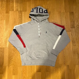 Grå hoodie från Polo Ralph Lauren - Säljer en grå hoodie från Polo Ralph Lauren med färgglada detaljer i rött, vitt och blått på ärmarna. Hoodien har en stor ficka framtill och justerbar dragsko i huvan. Perfekt för en avslappnad stil.