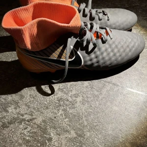 Grå och orange fotbollsskor från Nike - Snygga grå fotbollsskor med orange detaljer och snörning. Skorna har en hög krage för extra stöd och svarta dobbar för bra grepp på planen. Perfekta för fotbollsspelare som vill ha stil och funktion.