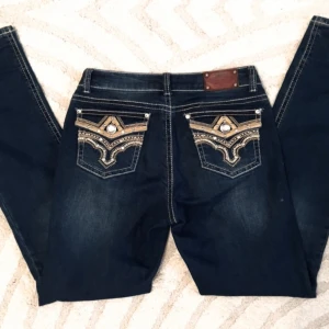 Mörkblå jeans med broderade detaljer  - Snygga mörkblå jeans från Stars & Stripes med unika broderade detaljer och glittriga stenar på bakfickorna. Perfekt för dig som vill sticka ut med stil. Jeansen har en klassisk femficksdesign och en bekväm passform. 38cm Midja 