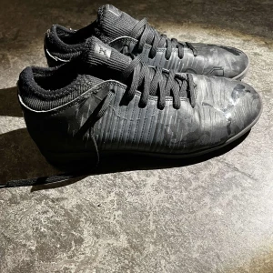 Svarta fotbollsskor med kamouflagemönster - Snygga svarta fotbollsskor med diskret kamouflagemönster och snörning. Perfekta för inomhusspel med en slitstark sula. Skorna har en modern design och är idealiska för den som vill ha stil på planen.