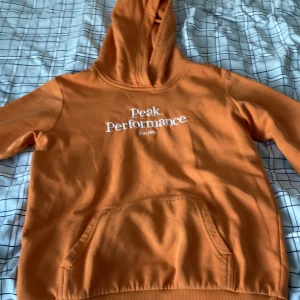 Orange hoodie från Peak Performance - Snygg orange hoodie från Peak Performance med vit logga på bröstet. Den har en klassisk känguruficka och en bekväm huva. Perfekt för en avslappnad stil.