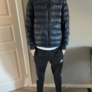 Svart dunjacka från Peak Performance - Säljer en stilren svart dunjacka från Peak Performance. Jackan har en dragkedja framtill och är långärmad, perfekt för kyligare dagar. Den är lätt och smidig att bära, vilket gör den idealisk för både stadsliv och naturäventyr.