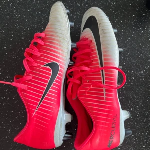 Nike Mercurial fotbollsskor - Säljer ett par Nike Mercurial fotbollsskor i röd och vit färg med svarta detaljer. Skorna har snörning och är designade för optimal prestanda på planen.