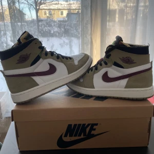 Nike Air Jordans 1 mid zoom air craft - Snygga Nike Air Jordans med en stilren design i beige och vinröd. Skorna har en hög modell med snörning och den klassiska Nike-loggan på sidan. Perfekta för dig som vill ha en sportig och trendig look.