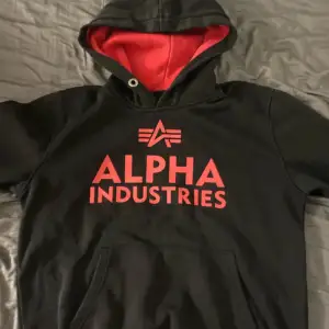 Säljer en svart hoodie från Alpha Industries med röd insida i huvan och röd logga på bröstet. Tröjan har en klassisk känguruficka och långa ärmar. Perfekt för en avslappnad stil.