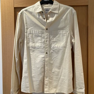 Beige långärmad skjorta från Zara - Snygg beige skjorta från Zara med långa ärmar och klassisk krage. Skjortan har två bröstfickor och knappar framtill. Perfekt för en stilren look.