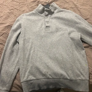 Grå tröja från Hugo Boss - Säljer en stilren grå tröja från Hugo Boss med hög krage och knappar. Tröjan har långa ärmar och ribbade muddar. Perfekt för en avslappnad och snygg look.