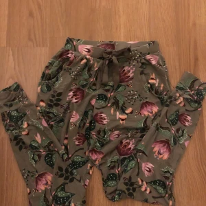 Blommiga byxor från Hunkemöller - Snygga och bekväma byxor från Hunkemöller med ett färgglatt blommönster i grönt och rosa. Byxorna har en avslappnad passform med snörning i midjan för extra komfort. Perfekta för en mysig dag hemma.
