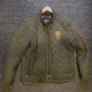 Grön quiltad jacka från Morris - Säljer en stilren grön quiltad jacka från Morris med broderad logga och brittisk flagga på bröstet. Jackan har dragkedja och långa ärmar, perfekt för kyligare dagar. Klassisk design med en touch av brittisk elegans. Passar L