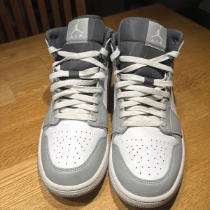 Nike Air Jordan 1 MID - Snygga Nike Air Jordans i en stilren kombination av grått och vitt. Skorna har klassisk snörning och en ikonisk design med Nike-loggan på sidan. Perfekta för dig som vill ha en sportig och trendig look. Storlek 43. Inköpta för drygt ett år sedan, använda en sommarsäsong. Använda med crease-skydd. I gott skick. 