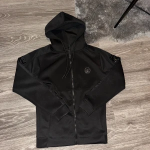 Svart hoodie från Icono Couture - Säljer en stilren svart hoodie från Icono Couture med dragkedja och huva. Tröjan har långa ärmar och ett märke på bröstet samt text längs ärmen. Perfekt för en casual look.