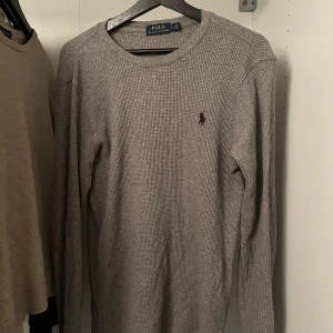 Grå tröja från Polo Ralph Lauren - Säljer en stilren grå tröja från Polo Ralph Lauren. Tröjan är tillverkad i bomull och har en diskret broderad logga på bröstet. Den har en klassisk rund hals och långa ärmar, perfekt för en avslappnad look.
