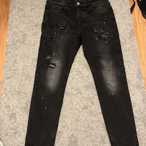 Svarta jeans med slitningar från Kings Will Dream - Snygga svarta jeans från Kings Will Dream med coola slitningar och färgstänk. De har en tight passform och är perfekta för en trendig look. Jeansen har klassisk femficksdesign och knappgylf.