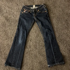 Mörkblåa true religion jeans med rosa detaljer - Snygga mörkblå bootcut jeans med unika detaljer på bakfickorna. Jeansen har en låg midja och klassisk femficksdesign med knappar och dragkedja framtill. Köpte Second hand i Thailand! ☺️ använts få gånger men har inga skavanker förutom märket på baksidan. pris kan diskuteras 🥰🥰🥰
