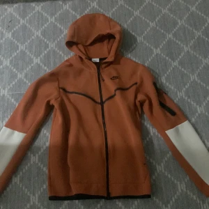 Orange hoodie från Nike - Säljer en snygg orange hoodie från Nike med dragkedja och huva. Den har långa ärmar med vita detaljer och en ficka på ärmen. Perfekt för en sportig look!
