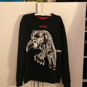 Svart långärmad tröja från Hugo Boss - Snygg svart långärmad tröja från Hugo Boss med ett stort tigertryck på framsidan. Tröjan har en rund halsringning och märket 'HUGO' i rött ovanför trycket. Perfekt för en cool och avslappnad stil.