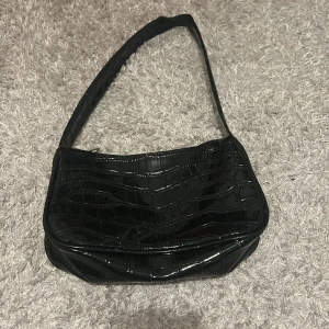 Svart axelväska i syntet - Snygg svart axelväska med krokodilpräglat mönster. Axelbandet har lite flisor, och det finns små skavanker. Men det är inget man märker om man inte tittar noga! 