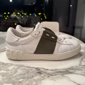 Trendiga Valentino Garavani Skor - Snygga vita Valentino Open sneakers med gröna detaljer och snörning. Skorna har en stilren design med en bred grön rem över snörningen och en dekorativ kant på sulan.