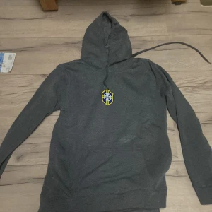 Grå hoodie med emblem - Säljer en grå hoodie med ett färgglatt emblem på bröstet. Tröjan har långa ärmar och en praktisk huva med dragsko. Perfekt för en avslappnad stil.