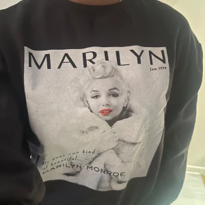 Svart/grå  tröja med Marilyn Monroe-tryck - Snygg svart tröja med ett stort tryck av Marilyn Monroe på framsidan. Perfekt för dig som vill ha en ikonisk look med en touch av retro. Tröjan har långa ärmar och en bekväm passform.