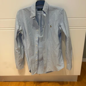 Ljusblå skjorta från Ralph Lauren - Snygg ljusblå skjorta från Ralph Lauren med klassisk krage och broderad logga på bröstet. Skjortan har långa ärmar och knappar framtill. Perfekt för en stilren look.