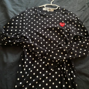 Svart prickig tröja från Comme des Garçons - Säljer en svart långärmad tröja från Comme des Garçons med vita prickar och det ikoniska röda hjärtat på bröstet. Perfekt för en stilren och lekfull look. Tröjan är i storlek M och har en normal passform. Pris går att diskutera vid snabb affär