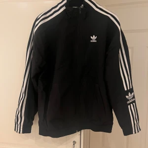 Windbreaker adidas originals - Säljer en klassisk svart träningsjacka från Adidas med de ikoniska tre vita ränderna längs ärmarna. Jackan har dragkedja framtill och Adidas-loggan på bröstet och ärmen. Perfekt för en sportig look! Varan är i nytt skick.
