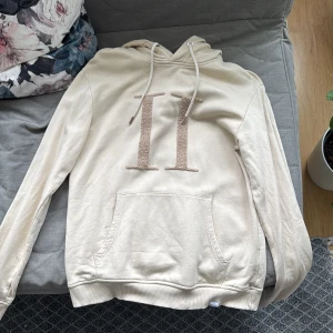 Beige hoodie med broderad detalj - Säljer en stilren beige hoodie med broderad detalj på framsidan. Tröjan har en klassisk design med dragsko i huvan och en stor ficka framtill. Perfekt för en avslappnad stil.