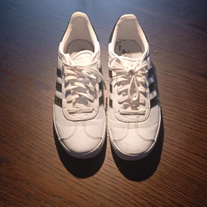 Vita Adidas Gazelle sneakers - Snygga vita Adidas Gazelle sneakers med klassiska svarta ränder. Skorna har snörning och en stilren design som passar perfekt till vardags. De är tillverkade i syntetmaterial och har en bekväm passform.Dem är bara använda ett fåtal gånger, dem är köpta i paris