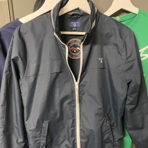 Mörkblå vindjacka från GANT - Snygg mörkblå vindjacka från GANT med dragkedja och broderad logga på bröstet. Jackan har en stilren design med hög krage och praktiska fickor. Perfekt för blåsiga dagar. Strl 158-164 13-14 år.