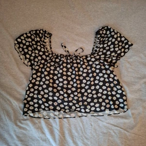 Svart blommig blus - Charmig svart blus med vita blommor och korta ärmar. Blusen har en söt knytning framtill och en luftig passform, perfekt för varma dagar.