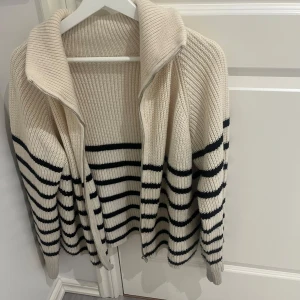 Beige och svart randig kofta - Säljer en mysig beige kofta med svarta ränder. Koftan har en dragkedja framtill och långa ärmar. Perfekt för kyligare dagar.