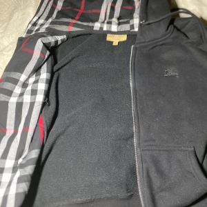 Svart zip up  från Burberry - Snygg svart hoodie från Burberry med dragkedja och klassiskt rutigt mönster i fodret. Den har en broderad logga på bröstet och en justerbar huva. Perfekt för en stilren look.