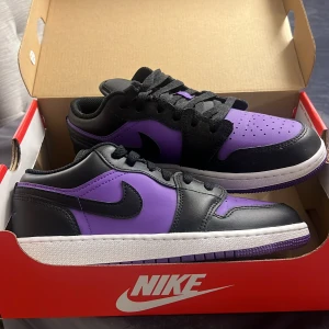 Nike Jordan 1 Low i svart och lila - Snygga Nike Jordan 1 Low i en cool kombination av svart och lila. Skorna har en klassisk design med snörning och en vit sula. Nyskick då de enbart är använda två gång. Storlek 37,5 och är svåra att få tag på då de inte säljs längre, inköpta från Nikes egna hemsida. Tillhörande kartong medföljer.
