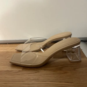 Beige sandaler med transparent klack - Stilrena beige sandaler med fyrkantig tå och transparent klack. De har en modern design med en klar rem över foten som ger en elegant look. Klacken är 6 cm