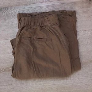 Bruna linnebyxor med resår i midjan - Säljer ett par bruna linnebyxor med resår i midjan och dragkedja. Byxorna har en avslappnad passform och är perfekta för en bekväm stil. De har också fickor på sidorna och bakfickor. 