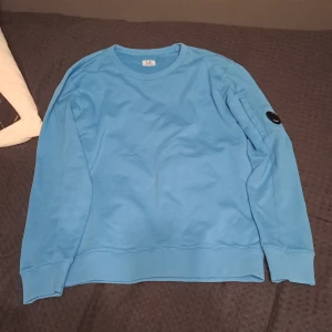 Blå tröja från C.P. Company - Säljer en snygg blå tröja från C.P. Company. Tröjan har långa ärmar och en rund halsringning. Den är dekorerad med en svart detalj på ärmen. Perfekt för en stilren look.