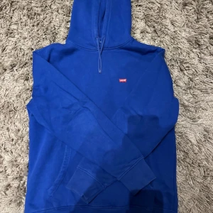 Blå hoodie från Levi's - Säljer en snygg blå hoodie från Levi's i storlek M. Den har en klassisk design med en liten röd Levi's-logga på bröstet och justerbar huva med dragsko. Perfekt för en avslappnad stil.