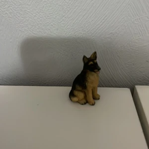 Leksaks hund - En liten dekorativ figur föreställande en hund, troligen en schäfer. Figuren är i naturliga färger med bruna och svarta nyanser. Perfekt som prydnad på hyllan eller skrivbordet.