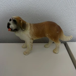 Bullyland hund - Charmig keramikfigur föreställande en hund med bruna och vita detaljer. Figuren har en realistisk design med en uttrycksfull ansiktsuttryck och en naturlig hållning. Perfekt som dekoration i hemmet eller som en present till hundälskare.