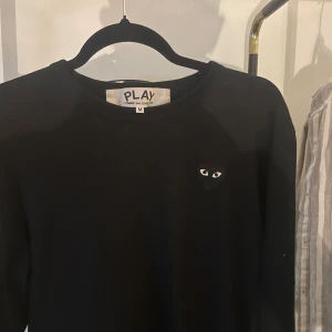 Svart tröja från Comme des Garçons - Säljer en stilren svart tröja från Comme des Garçons med det ikoniska hjärta med ögon på bröstet. Tröjan har en klassisk passform och är perfekt för en minimalistisk look. Passar bra till  jeans. Använd endast en gång, pris kan diskuteras vid snabb affär!!